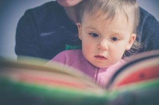 Toddler reading a book with their parent. Their parent is out of view and behind them.